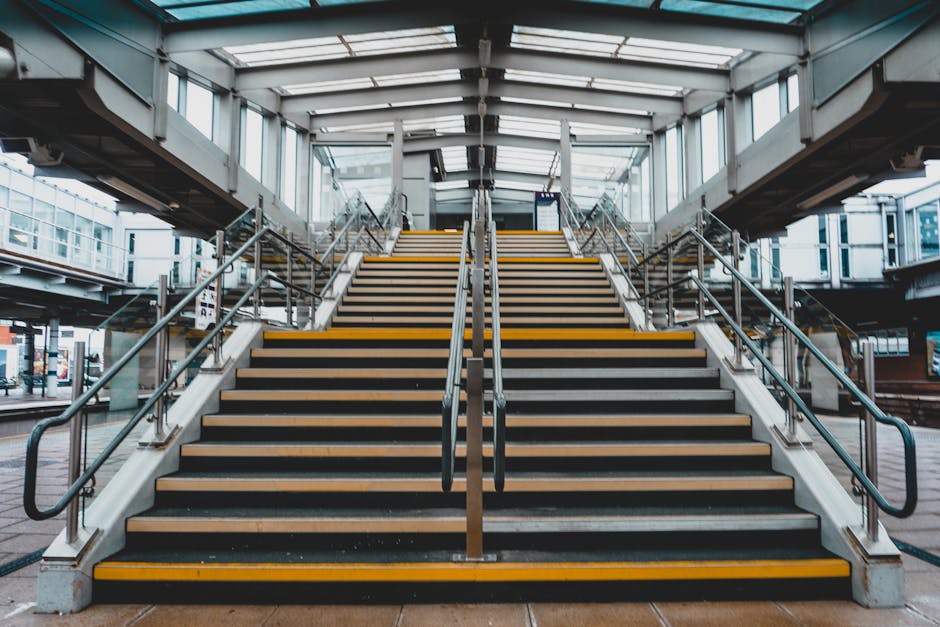 A wide view of an outdoor staircase at East Croydon station, leading up to a glass-covered footbridge. The staircase has black rubber treads with yellow safety strips along the edges, supported by met
