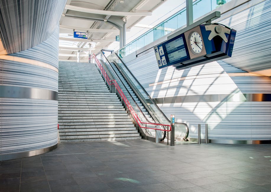 A wide view of an outdoor staircase at East Croydon station, leading up to a glass-covered footbridge. The staircase has black rubber treads with yellow safety strips along the edges, supported by met