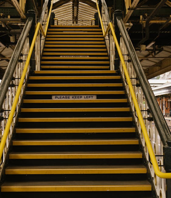 A set of industrial stairs with yellow handrails and black and yellow striped treads leading to an upper level inside a building, where a sign on one of the steps reads 'Please Keep Left.' The surroun