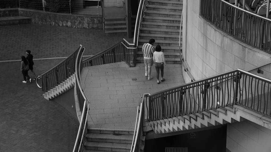 A set of industrial stairs with yellow handrails and black and yellow striped treads leading to an upper level inside a building, where a sign on one of the steps reads 'Please Keep Left.' The surroun